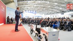 Ein Mann im blauen Anzug spricht auf einer Bühne in Halle B2 der transport logistic zu einem großen, sitzenden Publikum. Vor ihm steht ein E-Bike mit Anhänger.
