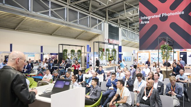Presentation at the Laser World of Photonics trade show in front of an audience, with exhibition booths and a sign reading “Integrated Photonics Area” in the background