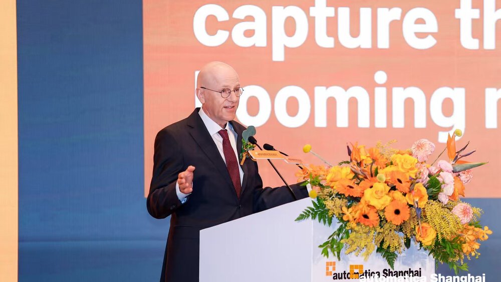 Man giving a presentation at a podium at automatica Shanghai with a slide and floral arrangement in front.