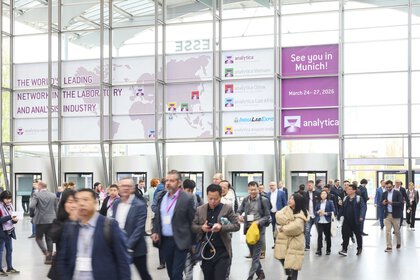 Trade fair visitors in the building at the West Entrance of Messe München. In the background there are glass walls and banners for the 'analytica' trade fair.