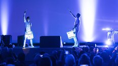 Zwei Schlagzeuger mit umgebundenen Trommeln stehen auf einer blau erleuchteten Bühne vor Publikum und zeigen eine Show.