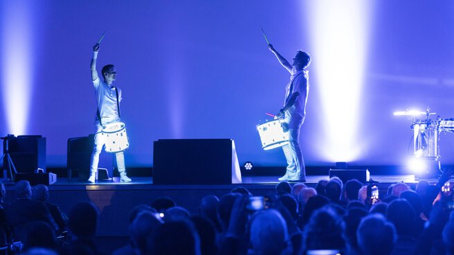 Two drummers with drums attached stand on a blue-lit stage in front of an audience and put on a show.