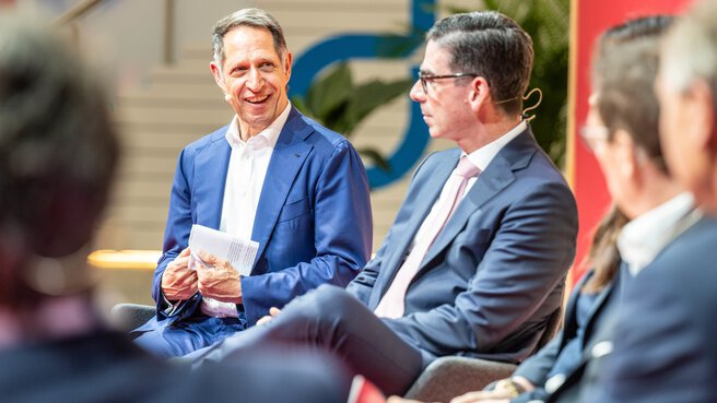 A gentleman in a blue suit sits on stage with other panellists during a panel discussion at the Transport and Logistic 2025 trade fair.