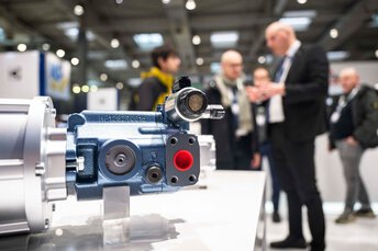 Industriebauteil auf einem Messestand im Fokus, Besucherinnen und Besucher unscharf im Hintergrund im Gespräch.