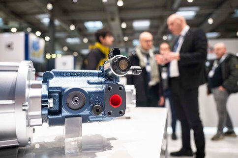 Industrial component displayed at a trade fair booth, with visitors blurred in the background in conversation.