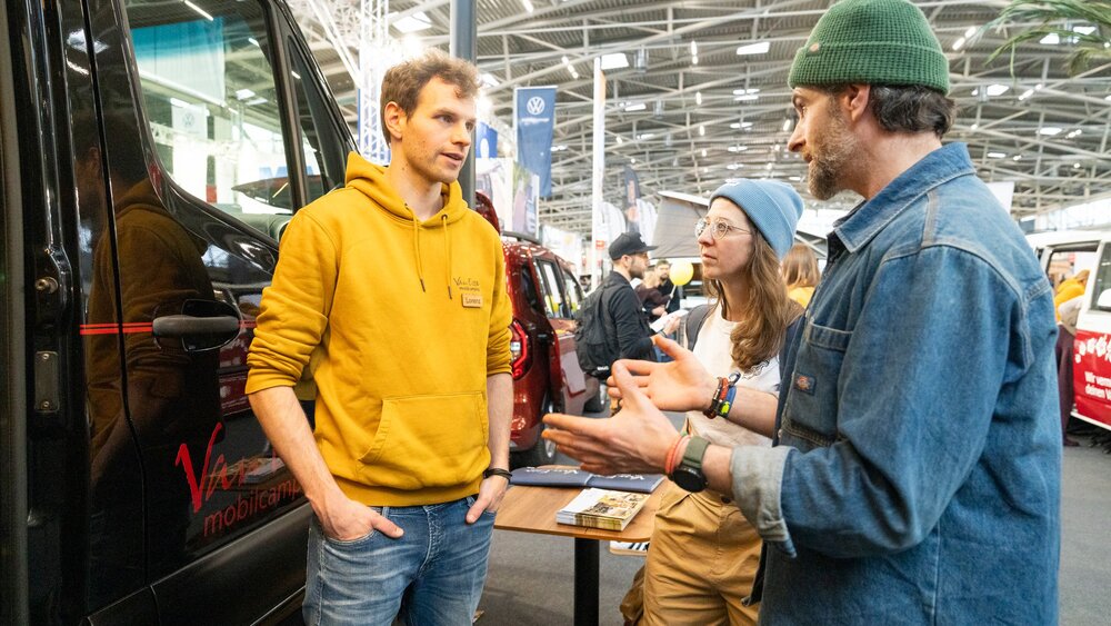 Drei Personen unterhalten sich bei einer Indoor-Veranstaltung neben einem Lieferwagen; auf einem kleinen Tisch daneben liegen Broschüren.