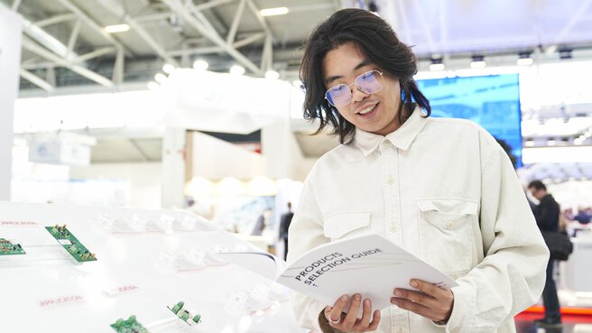 A man in a white corduroy shirt stands at an exhibition stand and reads a brochure with the inscription 