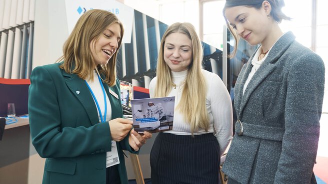Three young women stand at a trade fair booth and look together at a brochure held by one of them. They are smiling and appear engaged, with elements of the exhibition stand visible in the background.