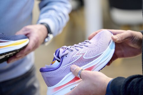 Nahaufnahme eines lila Sportschuhs mit weiß-rot-blauer Sohle, welcher von einer Person  in der Hand gehalten wird.