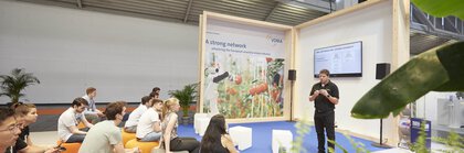 A speaker gives a presentation to a seated audience on a trade fair stage.