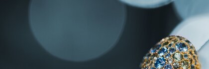 Nahaufnahme von einem Goldring mit blauen Edelsteinen in der rechten unteren Ecke und einem dunklen Hintergrund mit kreisförmigen Lichtreflexionen.