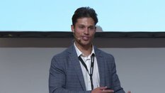 Eine Person im karierten Blazer und weißem Hemd spricht mit einem Mikrofon-Headset auf der Bühne.