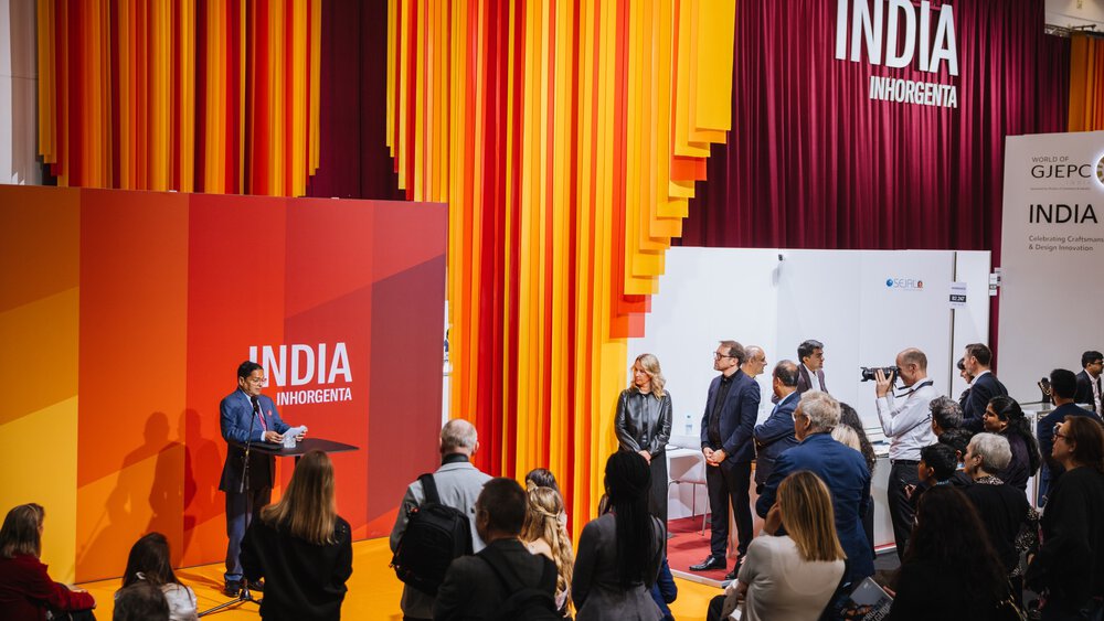 Person, die an einem Podium vor orangefarbenen und roten Vorhängen mit der Aufschrift „India Inhorigenta“ vor Publikum spricht.