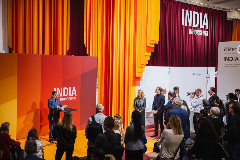 Person, die an einem Podium vor orangefarbenen und roten Vorhängen mit der Aufschrift „India Inhorigenta“ vor Publikum spricht.