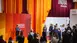 Person speaking to an audience at a podium in front of orange and red curtains with the inscription “India Inhorigenta”.