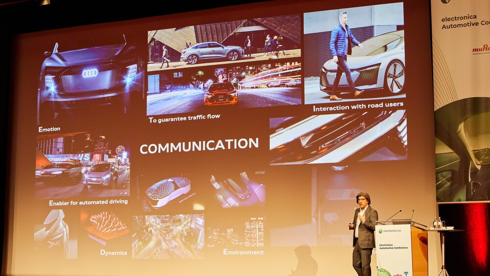 A man on the electronica stage stands next to the podium and speaks to the audience. Behind him, a presentation with the keyword 