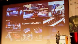A man on the electronica stage stands next to the podium and speaks to the audience. Behind him, a presentation with the keyword 