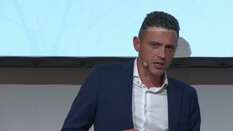 Ein Mann im blauen Anzug mit weißem Hemd spricht in einem Mikrofon-Headset vor einem hellen Hintergrund.