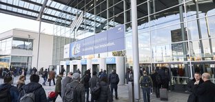 Zahlreiche Besucher betreten die Eingangshalle der productronica Messe durch Drehtüren. Fokus auf die Glasfassade des Eingangsbereich mit dem Messe München Logo.