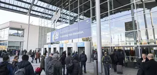 Numerous visitors enter the entrance hall of the productronica trade fair through revolving doors. Focus on the glass façade of the entrance area with the Messe München logo.