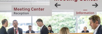 Besucherinnen und Besucher stehen im Eingangsbereich des Meeting Centers der EXPO REAL in München. Über ihnen hängen Schilder mit den Aufschriften „Meeting Center Reception“ und „Information“. Im Hintergrund sitzen Mitarbeitende hinter einem Empfangstresen, während mehrere Personen im Anzug im Vordergrund Gespräche führen oder auf ihre Tablets schauen.