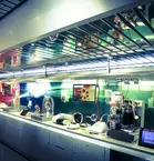 A modern laboratory with various scientific devices on a long work surface, illuminated by blue-green lights. Screens and mirrors can be seen in the background.