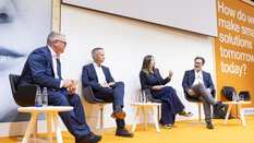 Four people in business attire sit on a stage with an orange floor during a panel discussion.