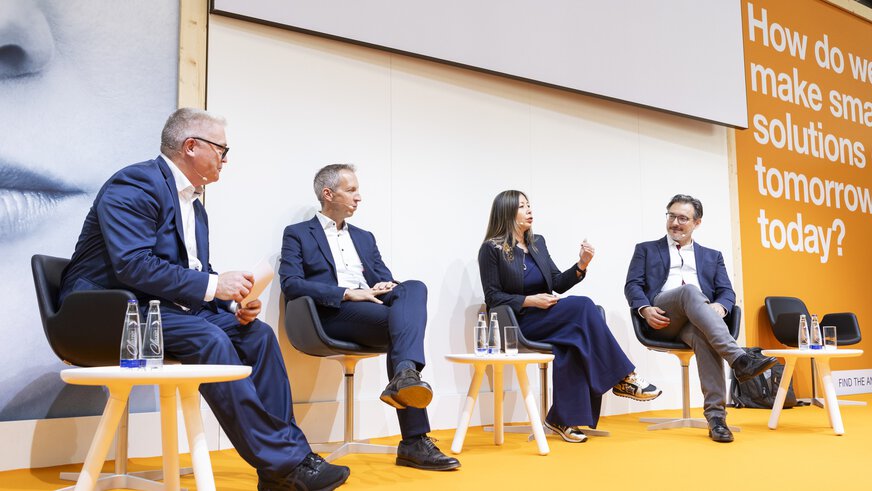Four people in business attire sit on a stage with an orange floor during a panel discussion.