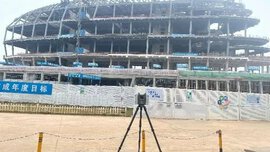 The shell of a curved modern building without an exterior façade, with a construction fence and a surveying instrument in front of it.