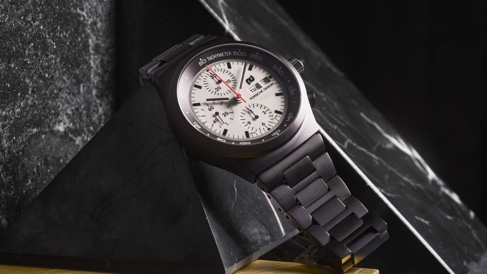 A black wristwatch with a white dial and chronograph is presented on a black marble pyramid.