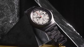 Eine schwarze Armbanduhr mit weißem Zifferblatt und Chronograph wird auf einer schwarzen Marmorpyramide präsentiert.