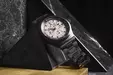 A black wristwatch with a white dial and chronograph is presented on a black marble pyramid.