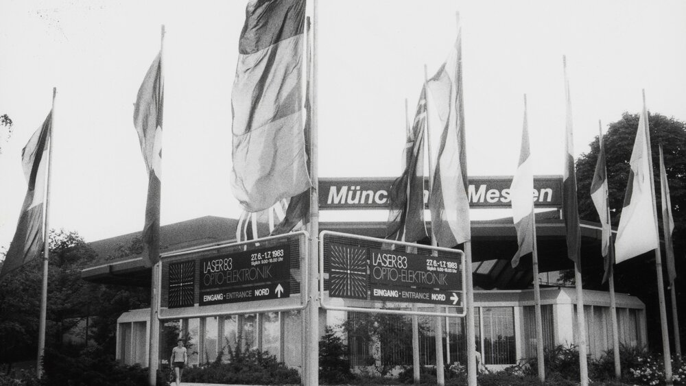 Black and white photo of flags and signs in front of the entrance to the LASER 83 trade fair.