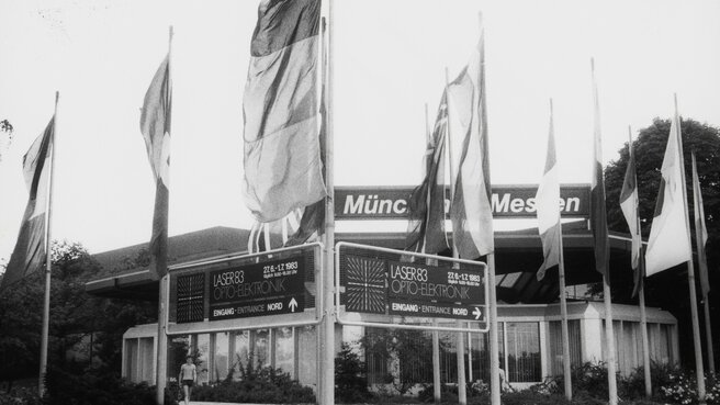 Black and white photo of flags and signs in front of the entrance to the LASER 83 trade fair.