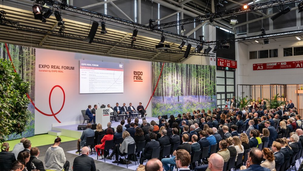 Numerous visitors sit in front of a podium at the EXPO REAL Forum and follow a discussion between several people in suits.