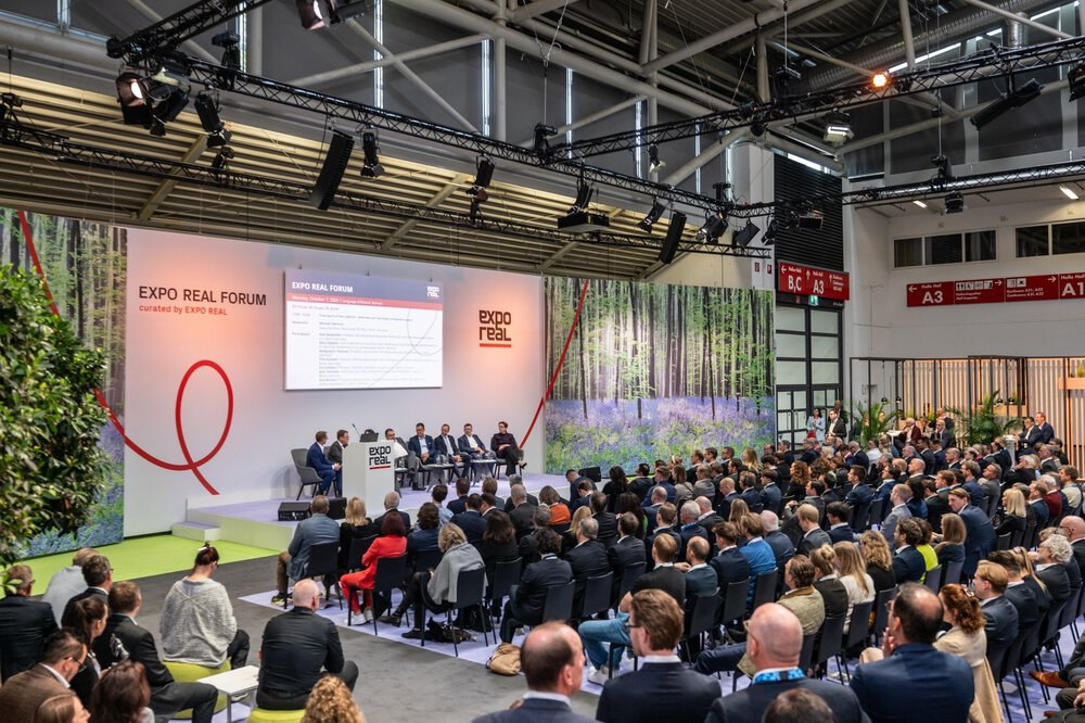 Numerous visitors sit in front of a podium at the EXPO REAL Forum and follow a discussion between several people in suits.