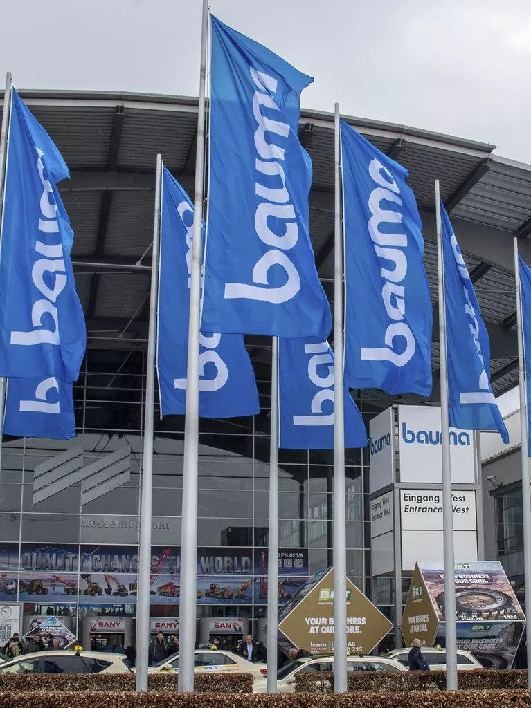 Bauma flags in front of a modern glass building, the entrance to the Messe München exhibition centre.
