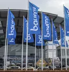 Bauma flags in front of a modern glass building, the entrance to the Messe München exhibition centre.