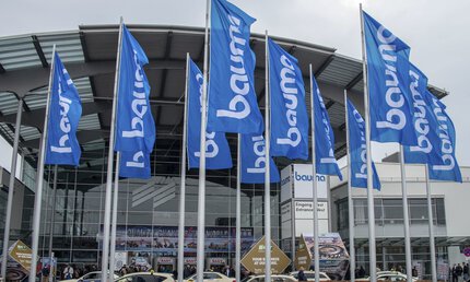 Bauma flags in front of a modern glass building, the entrance to the Messe München exhibition centre.