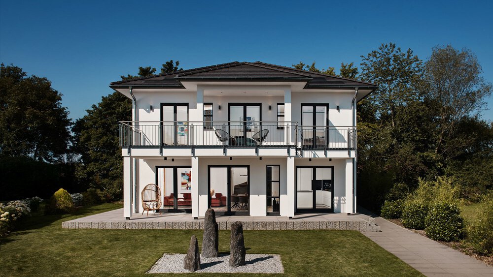 Modernes zweistöckiges weißes Haus mit großen Fenstern und Balkonen, umgeben von Grün unter einem klaren blauen Himmel.