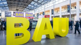 Große gelbe Buchstaben, die das Wort „BAU“ bilden, stehen im Eingangsbereich der „BAU 2025“. Besucher bewegen sich darum herum.
