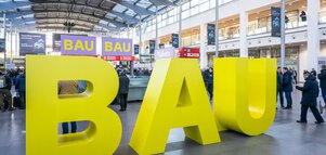 Große gelbe Buchstaben, die das Wort „BAU“ bilden, stehen im Eingangsbereich der „BAU 2025“. Besucher bewegen sich darum herum.