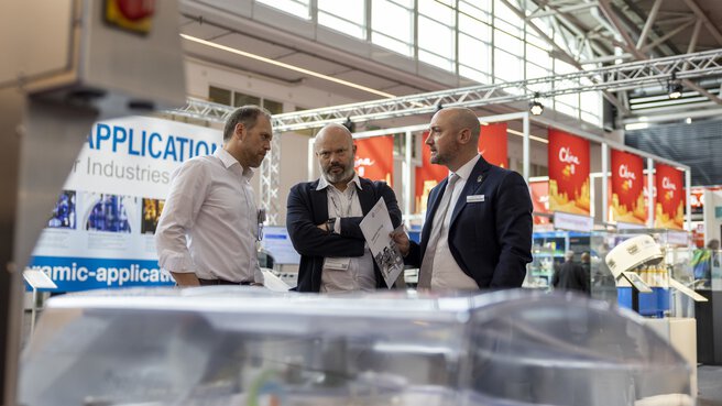 Drei Personen sprechen vor einem Messestand, im Hintergrund sieht man mehrere Austellungsstände.