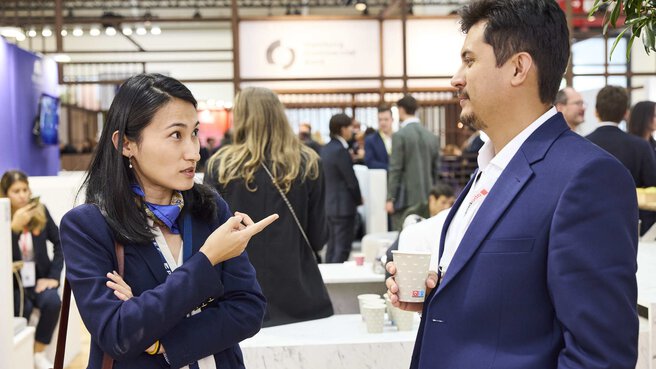 A woman gesticulates while talking to a man in a blue suit holding a paper cup in front of an exhibition stand.