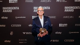 A smiling man with grey hair, glasses and wearing a suit stands in front of a photo wall of the INHORGENTA AWARD and holds a trophy.
