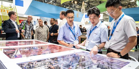 Three men look at an exhibit at a technology exhibition; various visitors and colorful stands can be seen in the background.