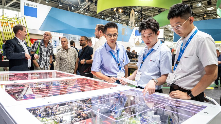 Drei Männer betrachten ein Exponat auf einer Technikausstellung, im Hintergrund sind verschiedene Besucher und bunte Stände zu sehen.