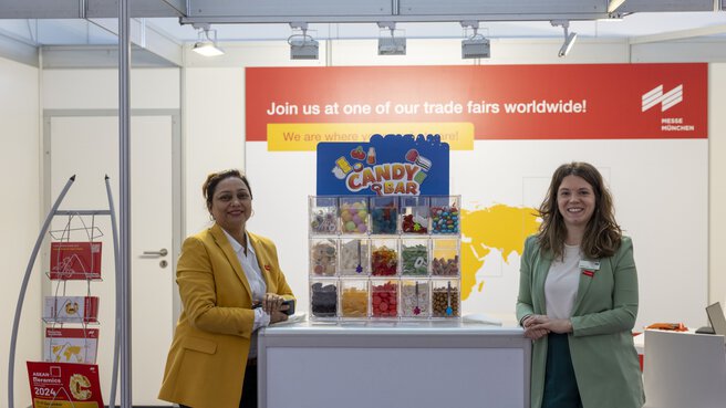 Zwei Frauen stehen lächelnd neben der Süßigkeitenbar am Messestand der ceramitec worldwide.