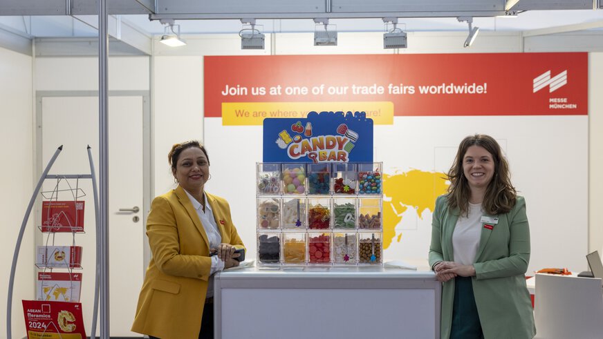 Zwei Frauen stehen lächelnd neben der Süßigkeitenbar am Messestand der ceramitec worldwide.