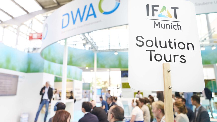 Hochgehaltenes Schild der IFAT Munich Solution Tours, dahinter sind unscharf einige Menschen zu sehen die einem Vortrag an einem Messestand lauschen.